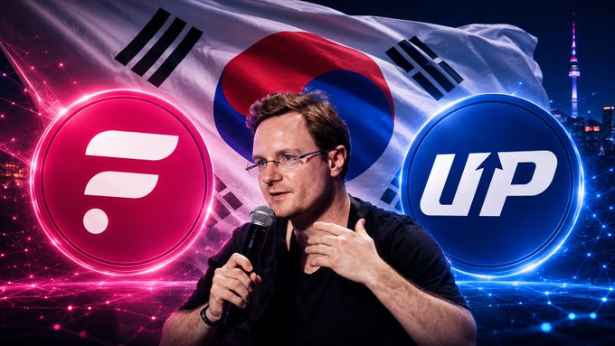 A man speaking into a microphone, with the South Korean flag in the background and two circular cryptocurrency logos flanking him: a pink logo and a blue 'UP' logo, set against a nighttime cityscape.
