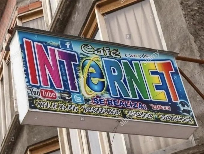 A storefront sign for 'Café Internet' advertising services like CD/DVD copying, transcriptions, prints, and investigations, with logos for Facebook, YouTube, Twitter, Google, and Yahoo.
