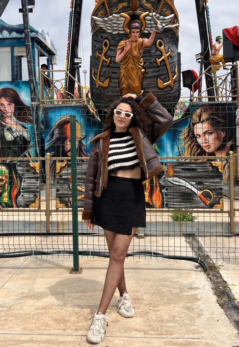 The image features a woman standing next to a ferris wheel at an amusement park. She is wearing a black skirt and jacket with white sneakers. The background of the photo shows various rides, including a pirate ship ride that has a statue of a man on top. The caption reads "GEvening Fam Half-Dead Not alive enough to remember spring, not dead enough to be free of pain", which seems to be related to the ferris wheel and its surroundings.