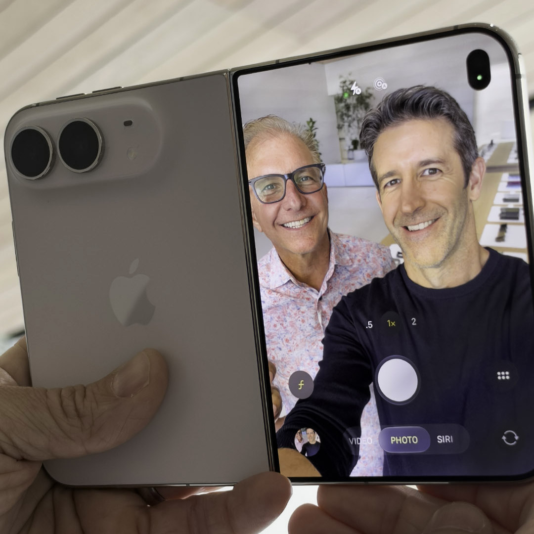 Two men taking a selfie video using a folded Apple iPhone, with the device's screen displaying the camera app interface and the rear showing its dual camera lenses.