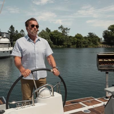 A man wearing sunglasses and a light blue shirt steers a boat on a calm body of water, with other boats and a tree-lined shore in the background.