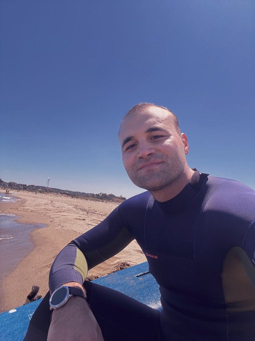 A man in a dark O'Neill wetsuit and smartwatch sits on a blue board on a sandy beach under a clear blue sky.