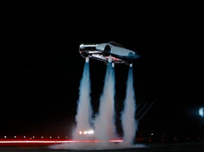 A Tesla Roadster hovers above the ground at night, propelled by four plumes of smoke/rocket exhaust, matching the post's depiction of the vehicle flying skyward. The image evokes the post's playful "Tesla to Mars" theme alongside the $TSLA ticker.