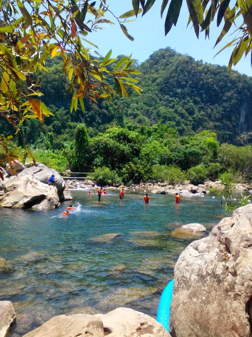 A scenic view of people wading in a clear river or natural pool surrounded by lush green mountains and large rocks, with a blue kayak visible in the foreground.