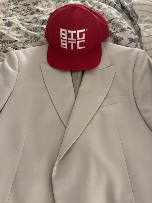 A red baseball cap with a white BTC/BTC logo resting on top of a cream-colored blazer, placed on a floral patterned bedspread.