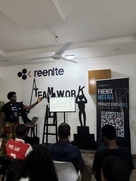 The image shows a group of people gathered around a presentation screen at an event. A woman stands behind the screen, giving a presentation or lecture about web3 privacy. The attendees are seated in chairs facing her, attentively listening and learning from her insights. There is also a potted plant visible in the background, adding some greenery to the room.