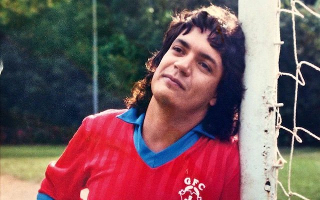 An older photograph of a young man with curly dark hair wearing a red soccer jersey with blue trim and a club crest, leaning against a white goal post net.