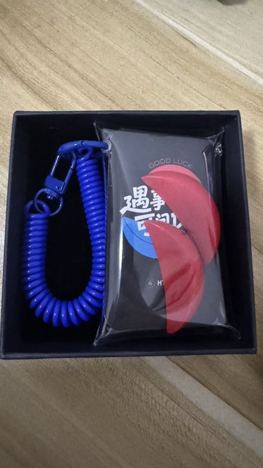 The image features a blue corded phone charm bracelet with a red charm, placed inside a black box or case. The charm bracelet has two charms - one red and one black. The charm bracelet itself is positioned against a wooden background, which adds an interesting contrast to its colors. The text in the image reads "Mike really loves Minnan culture!! Today'S Holy Grail toss: Asking if #BTC short position can make money! The result is a hint: Holy Grail!!! This time the merch really hits the spot for me @Xiaodu_HTX Contact now: Mike is arranging the same merch for".