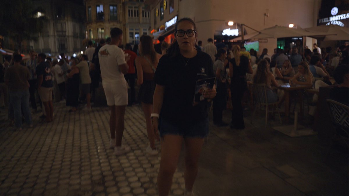 The image shows a woman walking down a street at night, holding a book or magazine. She appears to be enjoying her time outdoors as she walks past various people sitting outside of a bar. The scene captures the lively atmosphere of a city street during an event like the World Cup, with people gathered and socializing in front of shops and restaurants.