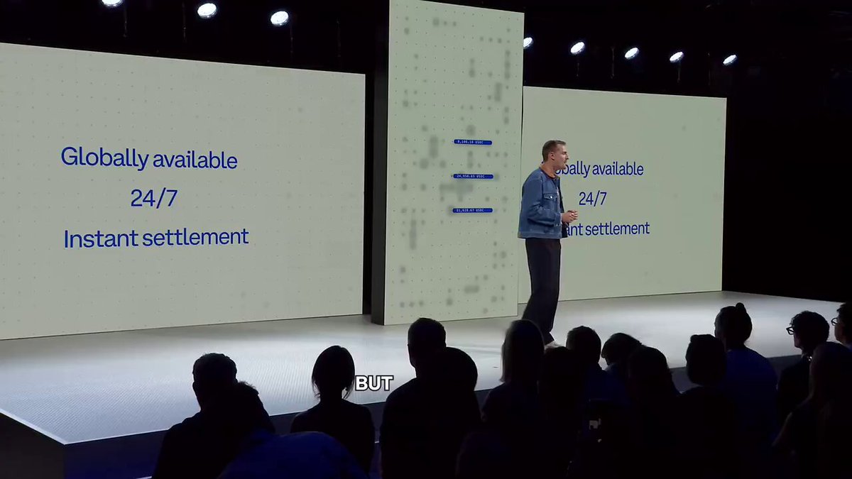 The image shows a man standing behind a podium at an event with a large screen displaying text about tokenization infrastructure. The man appears to be giving a speech or presentation related to this topic. The audience consists of several people, some sitting on chairs while others are standing around the stage. The setting is likely a conference or gathering where the speaker is addressing the importance of building native standards for stablecoins and other token-related projects.