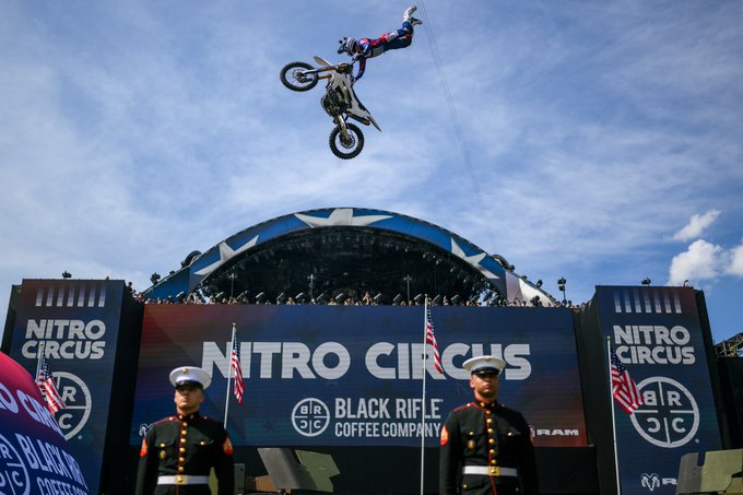 The image features a man performing a motorcycle stunt, flying through the air while riding his bike over a stage at an event called Nitro Circus. There are two men standing behind him, one wearing a military uniform and the other dressed as a cowboy. The stage is adorned with banners that say "Black Rifle Coffee Company" and "Kermit the Frog".