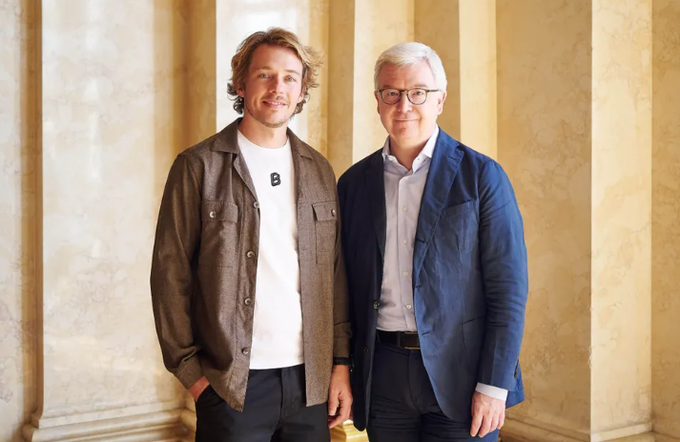 Two men posing for a portrait in an ornate hallway with marble columns; one wears a brown jacket with a Bitcoin logo shirt, and the other wears a blue blazer with glasses.