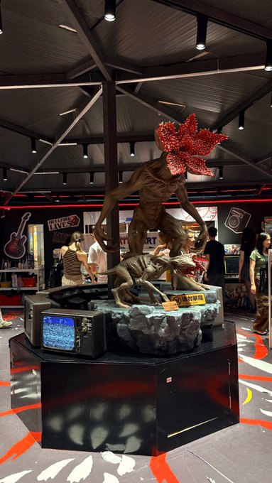 A large statue of a Demogorgon from Stranger Things with its head open stands on a rock-themed display inside what appears to be a Stranger Things merchandise store, with people browsing in the background.