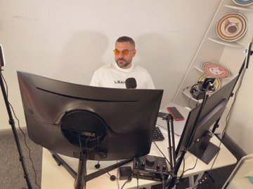A man in a white "USA" shirt sits in a recording studio setting behind a large monitor with microphones and speakers visible, streaming or hosting a discussion likely about cryptocurrency topics mentioned in the accompanying post.
