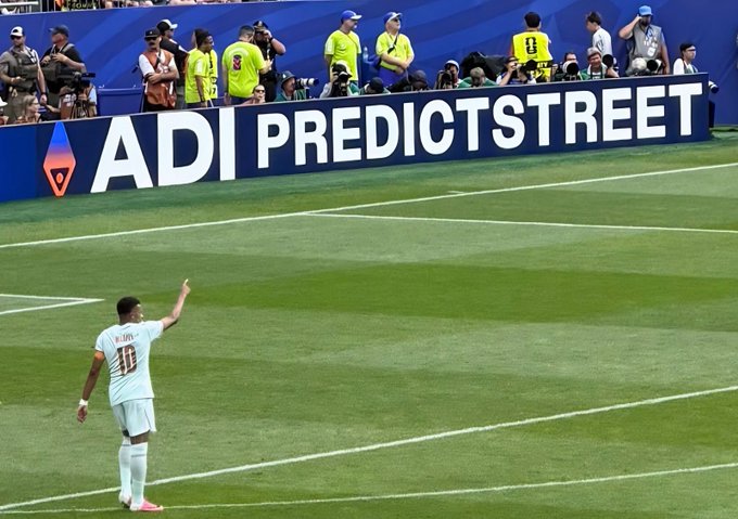 A soccer player on a field raising his arm in celebration, with an advertising board reading 'ADI PREDICTSTREET' and photographers visible in the background.
