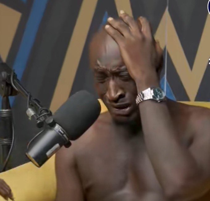 A shirtless man sitting in front of a podcast microphone with his hand on his head, wearing a silver watch, with a shocked or distressed expression on his face.