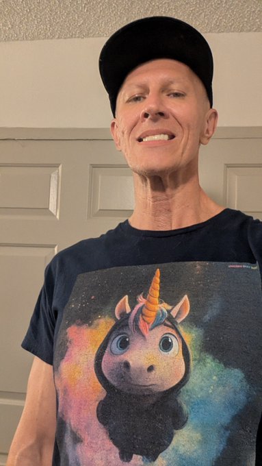 A selfie of a smiling man wearing a black cap and a dark t-shirt featuring a colorful graphic of a unicorn.