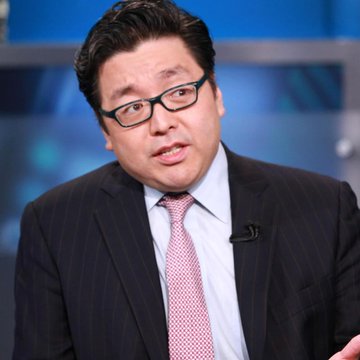 A man wearing glasses, a dark pinstripe suit, and a pink tie is speaking during what appears to be a television interview, likely a news segment about Tom Lee.
