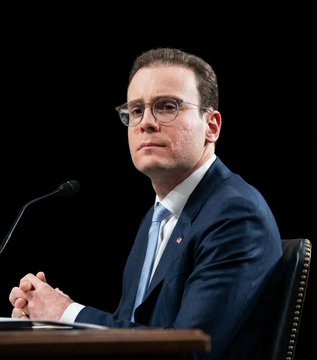 In this image, a man wearing glasses is sitting at a desk with his hands clasped together, looking directly at the camera. The setting appears to be an indoor room or office environment, as there are chairs present around him. The main focus of the image is on the man and his attire, which includes a suit and tie.