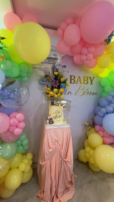 A pastel-themed "BABY in Bloom" baby shower setup featuring a cake decorated with "Oh Baby" on a draped peach table, framed by an arch of pink, yellow, green, and blue balloons and floral arrangements. The scene reflects the post's mention of planning a surprise baby shower, capturing the festive, celebratory atmosphere of the event.