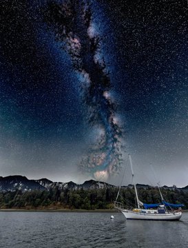 A stunning night sky photograph showing the Milky Way galaxy stretching across the sky above a lake with a white sailboat anchored near forested mountains.