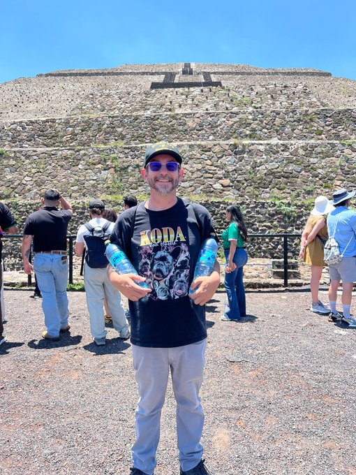 The image shows a man standing next to a large stone wall with his arms raised above his head, holding two water bottles. The man appears to be posing for a photo at the Pyramids of Teotihuacan. In the background, there are other people present as well. The caption on the post suggests that visitors should wear an @BoredApeYC hat when visiting this historical site.