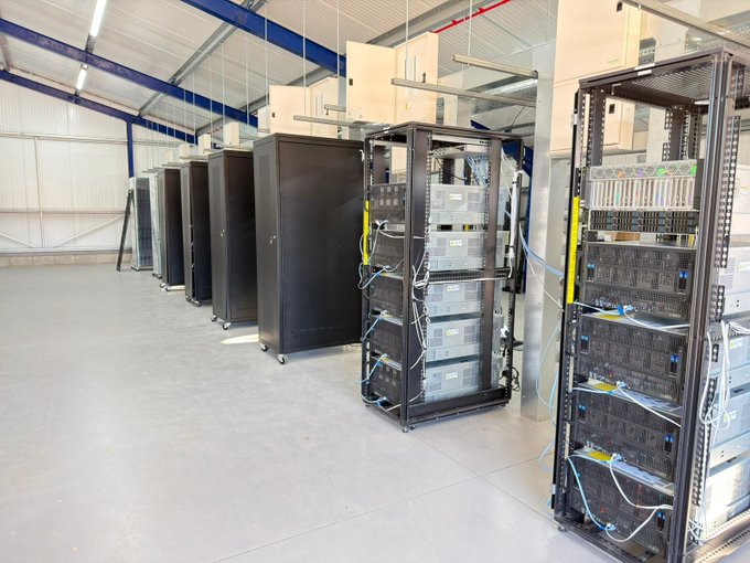 A row of black server racks and networking equipment lined up in a data center or industrial facility with white walls and ceiling beams.
