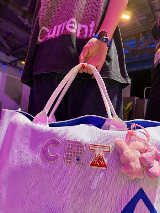 A close-up of a person wearing a black t-shirt with 'Current' printed on it, holding a white tote bag featuring the word 'CRZY' in colorful patches and a small pink plush bunny keychain attached to the handle.