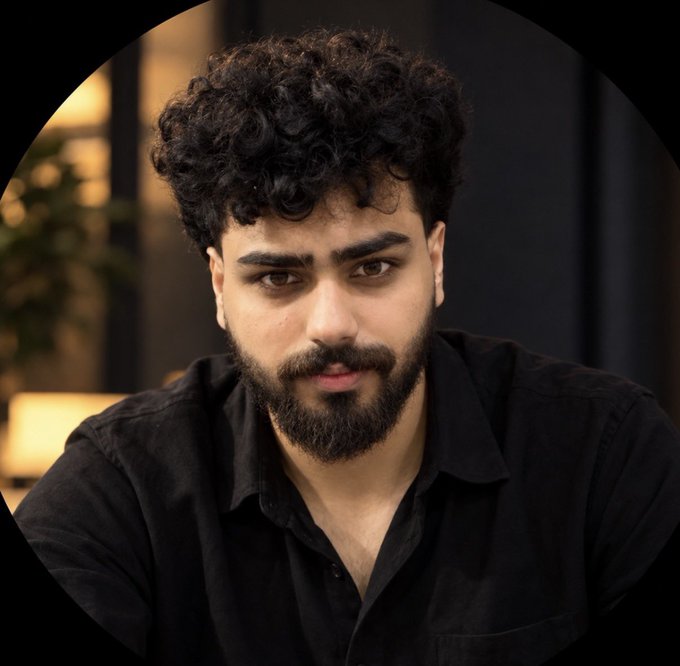 A portrait of a man with curly dark hair and a beard, wearing a black collared shirt, looking directly at the camera.
