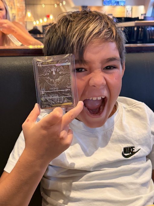 A young boy excitedly displays a holographic Pokémon trading card, wearing a white Nike t-shirt with his mouth wide open in a joyous shout, perfectly capturing the "#Cardano fam" excitement over pulling a favorite card.