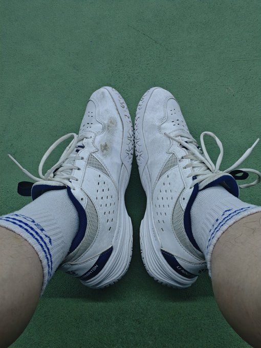 A pair of white and blue athletic shoes worn with white socks on a green surface, showing visible dirt stains on the toe area.
