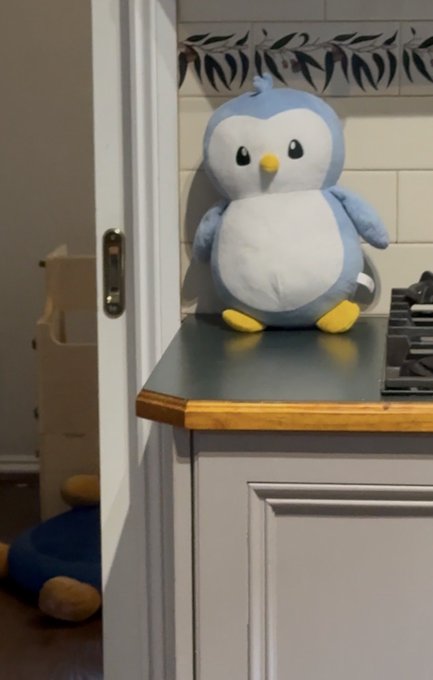 A blue and white penguin plush toy sitting on a kitchen counter, with another plush partially visible on the floor behind a doorway.