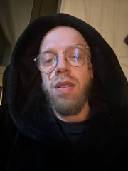 A close-up photo of a bearded man wearing round glasses and a dark hooded sweatshirt, appearing to be speaking or reacting.