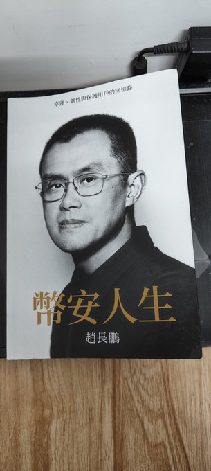 A book titled "幣安人生" by 趙長鵬 (CZ), founder of Binance, rests on a wooden surface. The cover features a portrait of Zhao in glasses and the subtitle reads "華語·新時代幣圈用戶的必讀書" (A must-read for Chinese-speaking users in the new era of crypto). The post expresses gratitude to a teacher named @Supers6061 for sending the book as a gift.