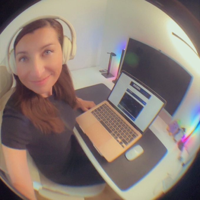 A fisheye lens photo of a smiling woman wearing white headphones at a desk with a laptop and an external monitor displaying Bloomberg content.