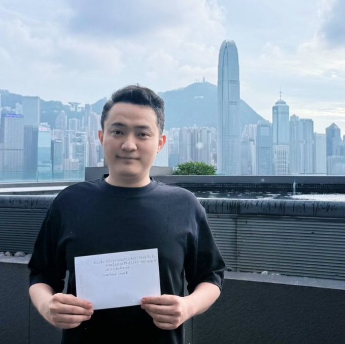 A young man in a black shirt holds up a handwritten note while standing on a rooftop with the Hong Kong skyline visible behind him.