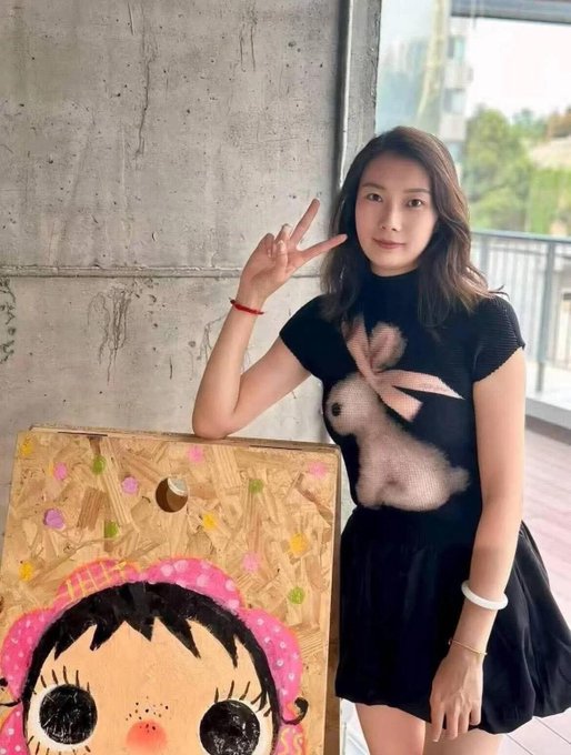A young woman wearing a black mesh top with a printed design and a white bangle, making a peace sign while standing next to a wooden board painted with a cartoon character.
