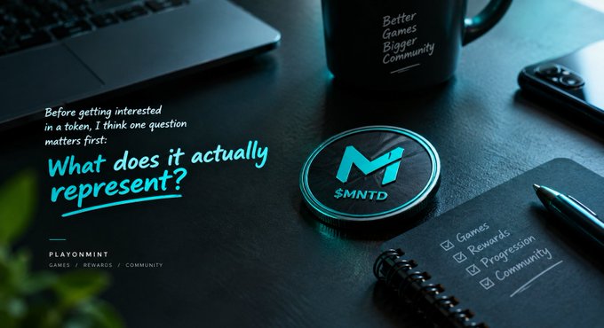 A desk setup featuring an '$MNTD' branded coaster with a cyan 'M' logo, a mug reading 'Better Games Bigger Community', and a checklist notebook next to a laptop.