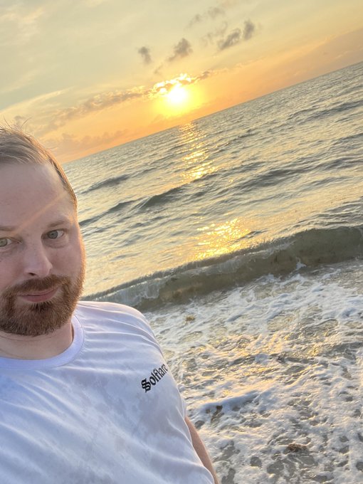 A man with a beard wearing a white t-shirt with the text "Solita" takes a selfie on a beach during sunset. The golden sun reflects brightly off the ocean waves behind him.