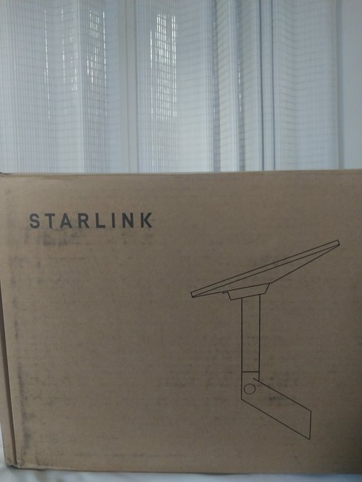 A cardboard box labeled 'STARLINK' with a line drawing of a satellite dish mount sits in front of sheer white curtains.