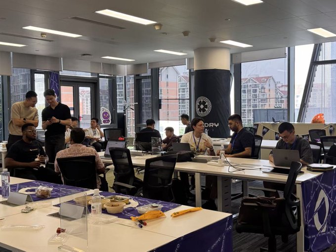 The image shows a group of people sitting at desks with laptops open, engaged in various activities such as working or chatting with each other. The room has white walls and large windows that provide ample natural light. There are several chairs placed around the room, some occupied by individuals focused on their work while others appear to be socializing.

The main focus of the image is a group of people sitting at desks in an office setting, surrounded by laptops and other computer equipment. The atmosphere seems collaborative and conducive to productivity.