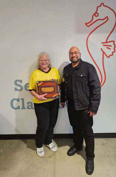 A woman in a yellow jersey holds an 'Ollie's Station' plaque next to a man in a black jacket, standing in front of a seahorse logo.