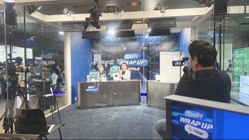 A behind-the-scenes view of a news studio showing camera equipment, a monitor displaying 'WRAP UP', and crew members working.