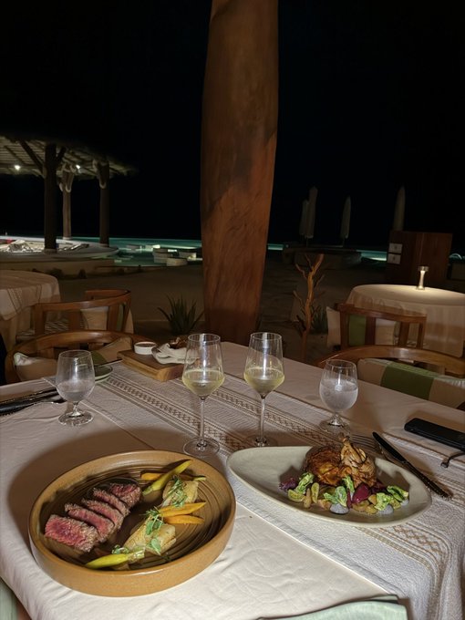 An upscale beachside dinner features a beautifully plated meal of seared steak with vegetables alongside a gourmet dish, accompanied by wine glasses on a candlelit table under the night sky. The elegant setting and thoughtful presentation reflect the post's sentiment of appreciating those who consistently show up and support in return.