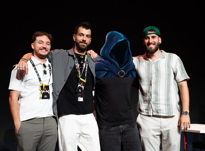 Three men pose at an event alongside a person in a black shirt and a blue hood featuring a golden ring, appearing as the 'Anon' character.