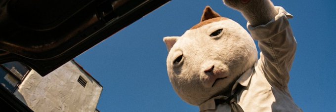 A person wearing an oversized cat head mask peers down into what appears to be the trunk of a car, captured from a low angle against a clear blue sky. The unusual mascot-like imagery mirrors the memecoin post's message about $KET being a different kind of "cat" investment—not a lottery ticket, but one worth watching closely and staying invested in.
