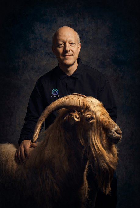 A professional portrait of a bald man wearing a dark polo shirt with a 'CIRCLE' logo, standing behind a large goat with prominent curved horns.