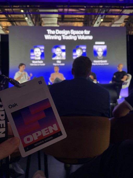 A view of a conference panel titled 'The Design Space for Winning Trading Volume' with speakers on stage, while an attendee in the foreground holds an 'Oak' badge labeled 'OPEN' with 'Reserve Access.'