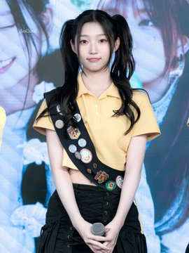 A young woman with pigtails stands on a stage wearing a yellow polo shirt and a black sash decorated with various colorful pins, holding a microphone.