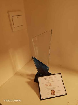 An award displayed in a clear acrylic case on a shelf, accompanied by a 'CERTIFICATE' card, shot on a Meizu 20 Pro.